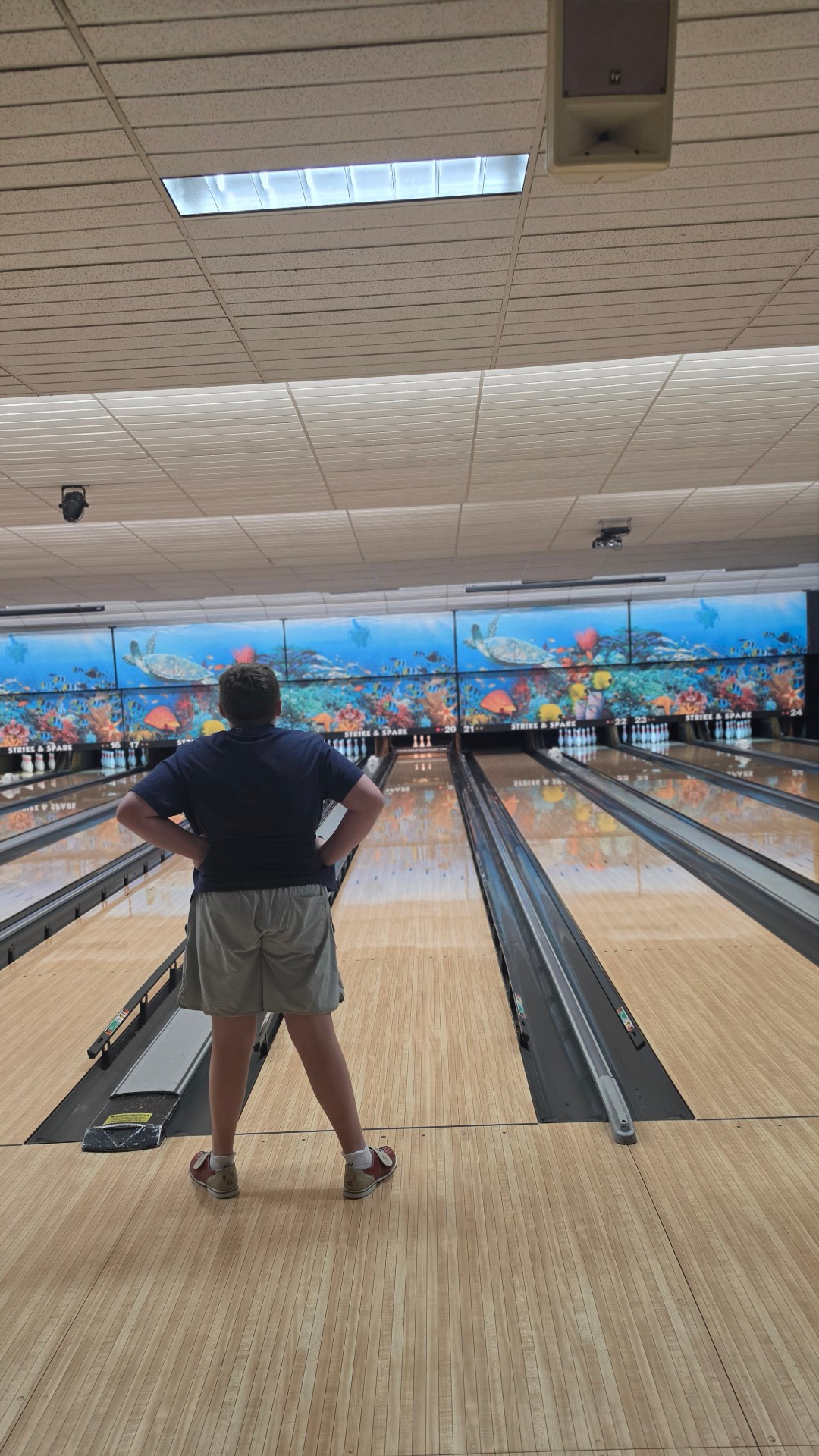A young person stands facing the bowling lanes and a colorful ocean mural.