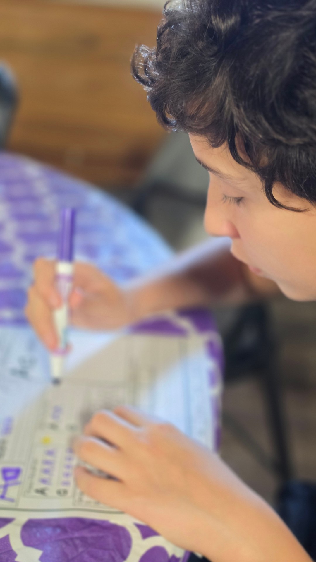 A learner writes on a worksheet with a purple pen at a patterned table.