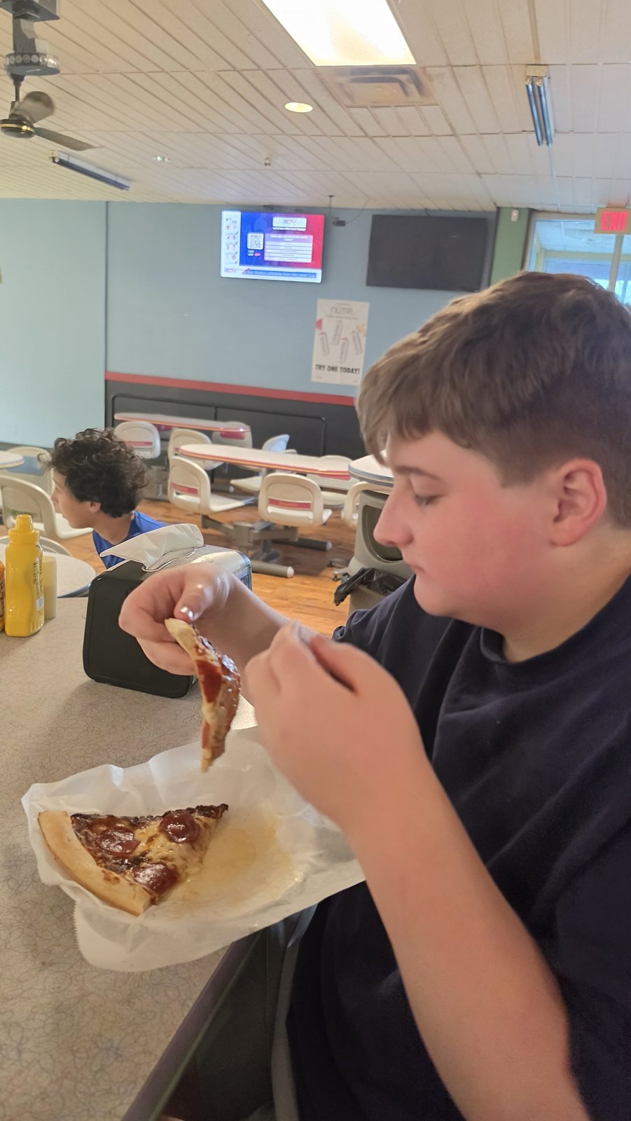 A young person enjoys a slice of pizza at the bowling alley counter.