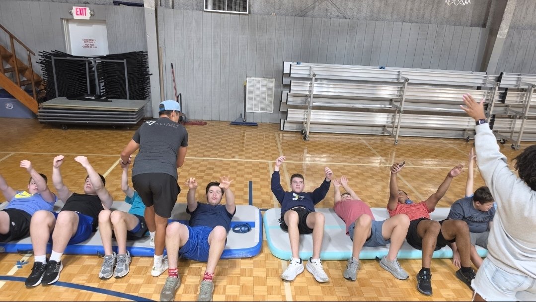 The group does a core exercise on mats in the gym with a coach guiding them.