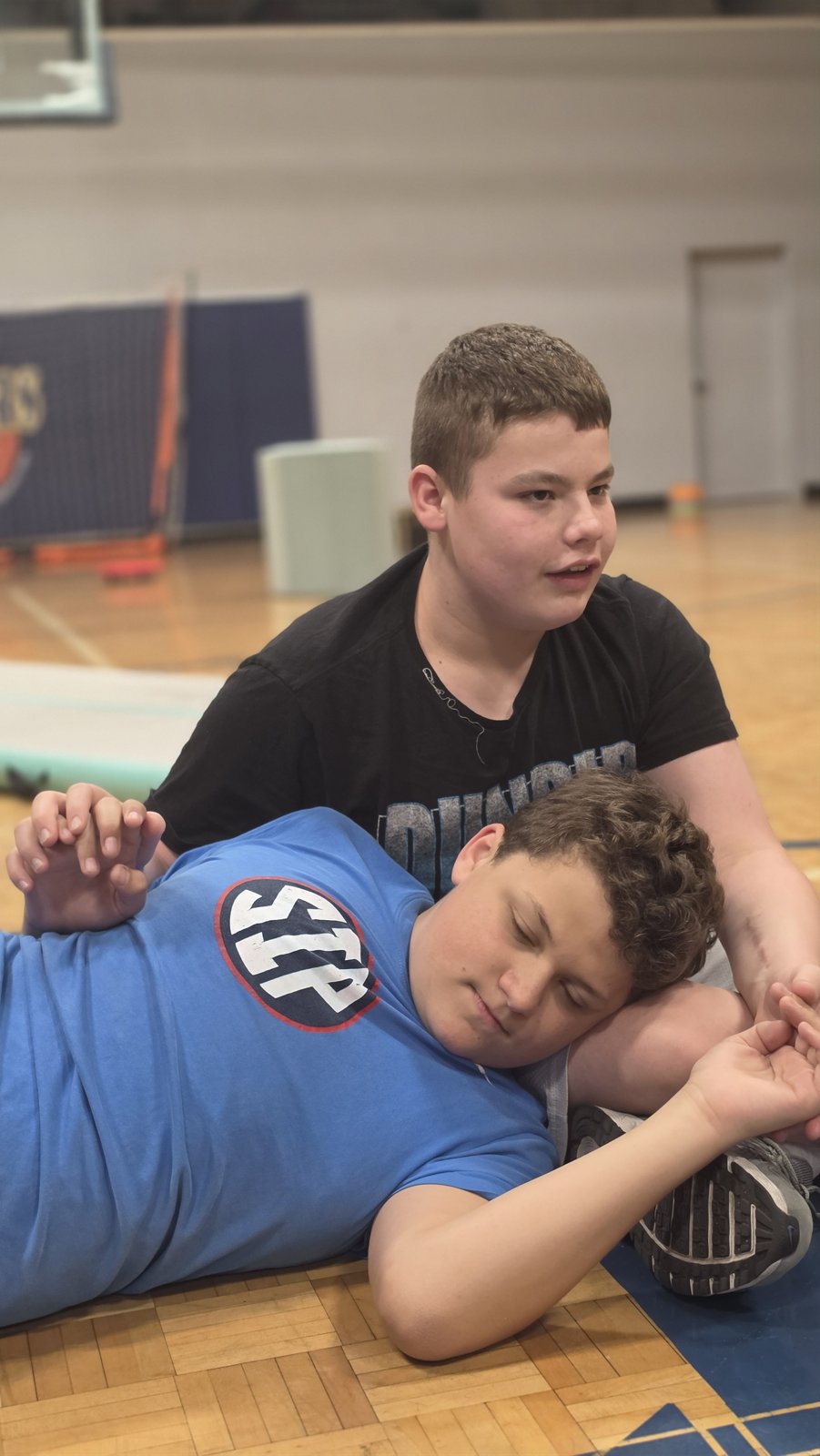 Two young people take a break on the gym floor, one resting their head on the other's lap.