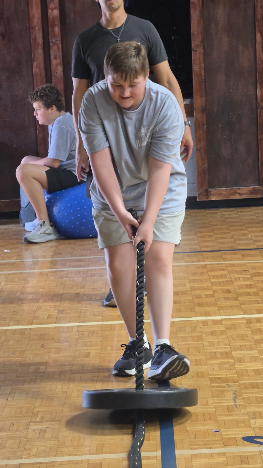 A young person pulls a weighted rope across the gym floor with an adult standing behind them.