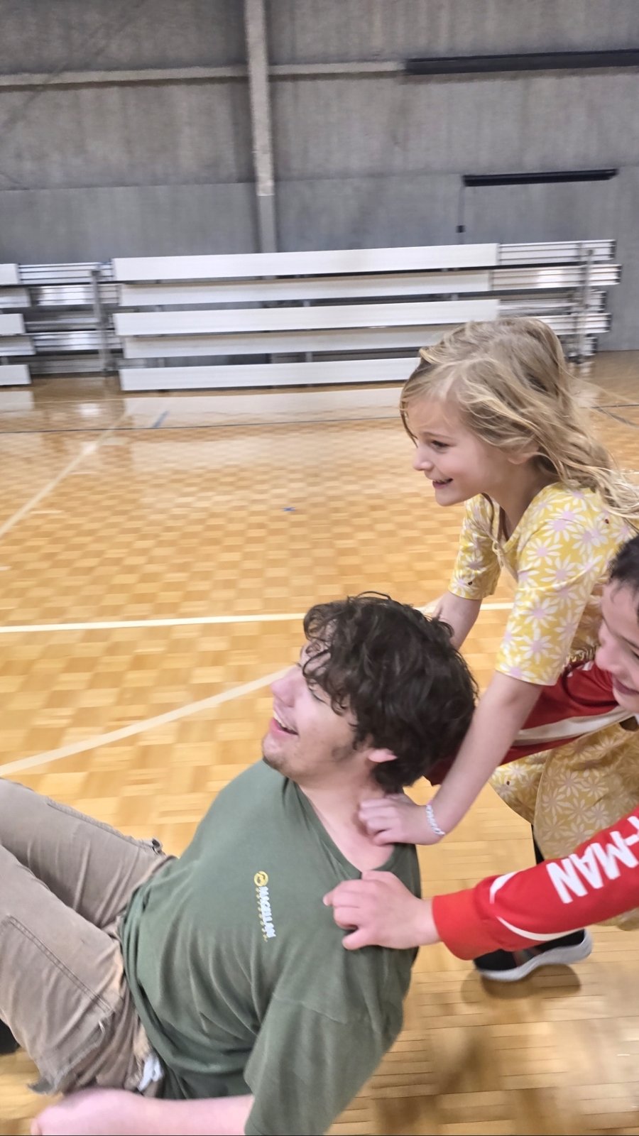 Kids playfully pile onto a laughing staff member on the gym floor.