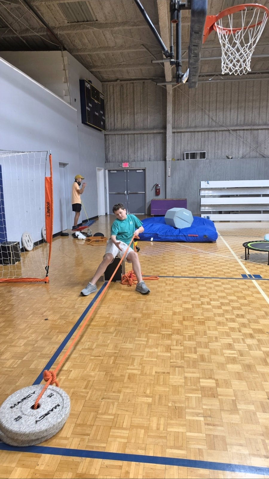 A boy pulls a rope attached to a weight plate across the gym floor.