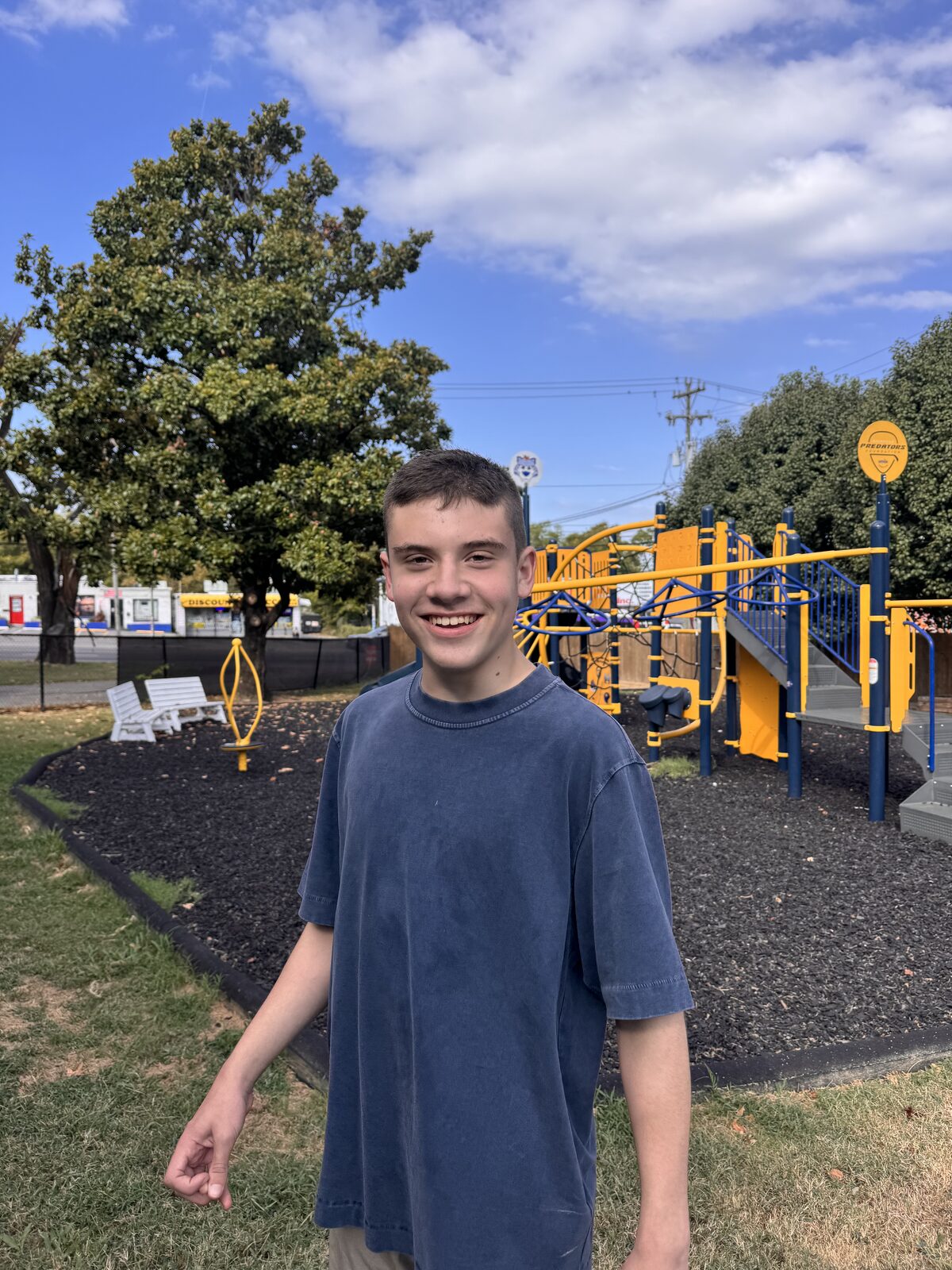 A smiling teen stands outdoors in front of a playground on a sunny day.