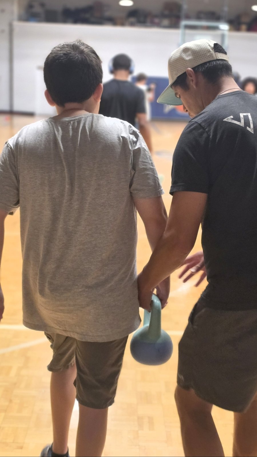 A coach guides a young athlete holding a kettlebell on the gym floor.