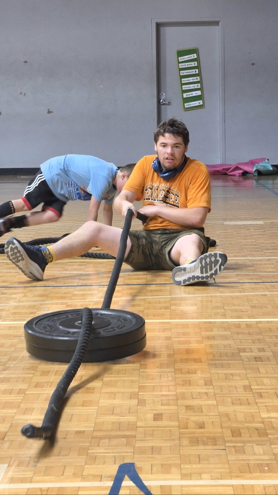 A young man pulls a weighted sled with a rope across the gym floor while a teammate works out behind him.