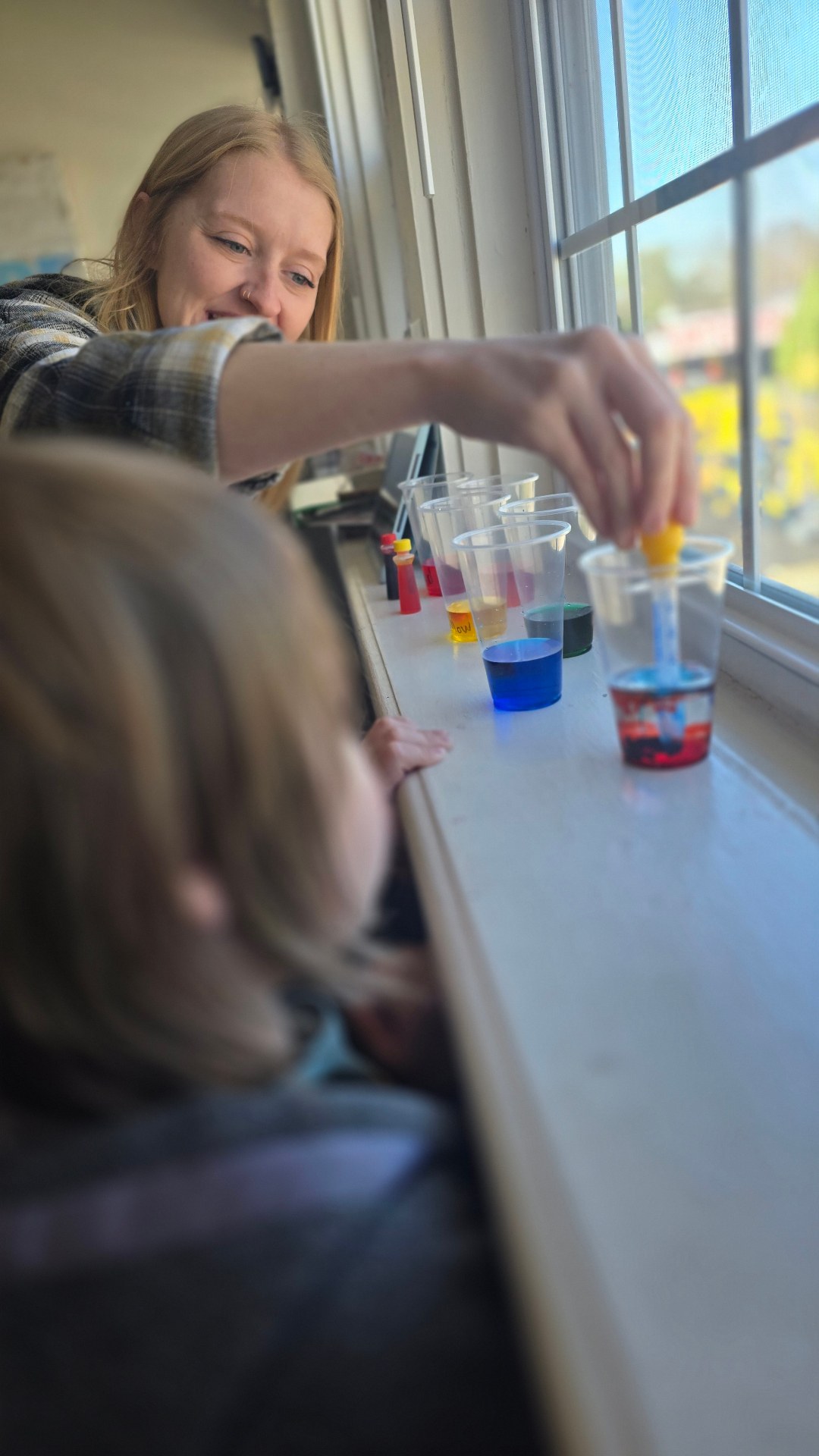 An adult mixes colored water at a window ledge while a child watches.