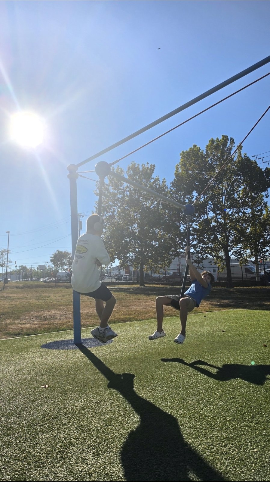 Two kids ride a playground zip-line together in the sunshine.