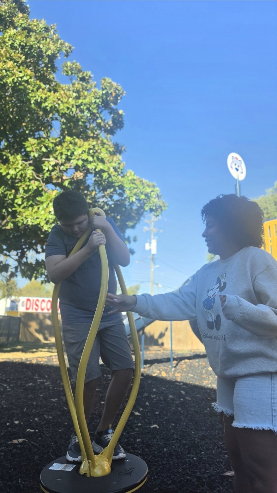 A boy rides a playground spinner while a friend spots him.
